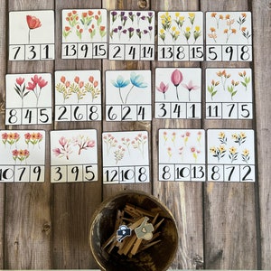 Può includere: Un insieme di flashcard a tema floreale con numeri, disposte su una superficie di legno. Ogni carta presenta un'illustrazione floreale ad acquerello e una serie di numeri. Una piccola ciotola con mollette di legno è in basso.