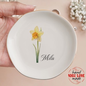 May include: A white ceramic trinket dish with a gold rim, featuring a watercolor daffodil and the name "Mila" in script. The dish is hand-made and perfect for holding jewelry or small items.