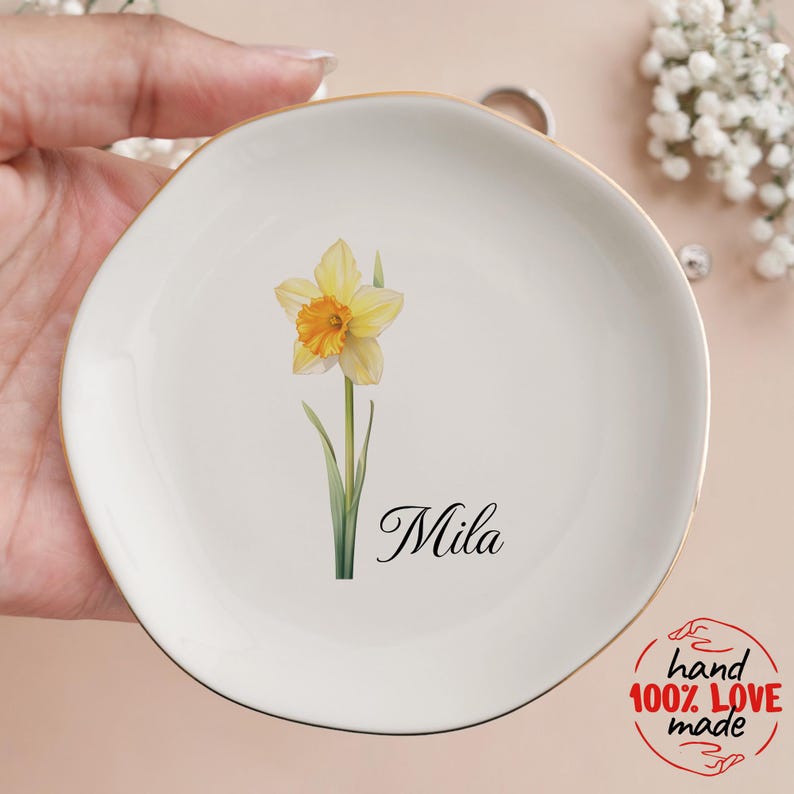 May include: A white ceramic trinket dish with a gold rim, featuring a watercolor daffodil and the name "Mila" in script. The dish is hand-made and perfect for holding jewelry or small items.