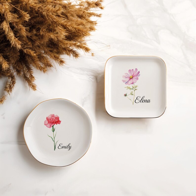 May include: Two white ceramic plates with gold rims. One is round with a red flower and the name "Emily". The other is square with a pink flower and the name "Elena". Decorative floral design.