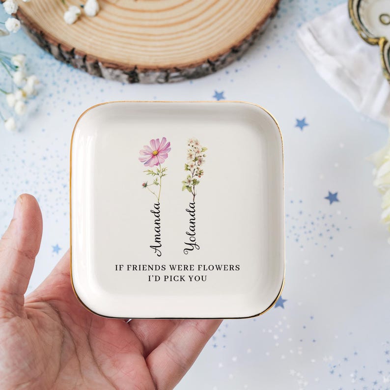 Pu&ograve; includere: Un piatto portaoggetti in ceramica bianca con bordo dorato. Il piatto presenta due fiori stilizzati con i nomi "Amanda" e "Yolanda" scritti verticalmente accanto a loro. Il testo "Se gli amici fossero fiori, ti sceglierei" &egrave; stampato sotto i fiori.