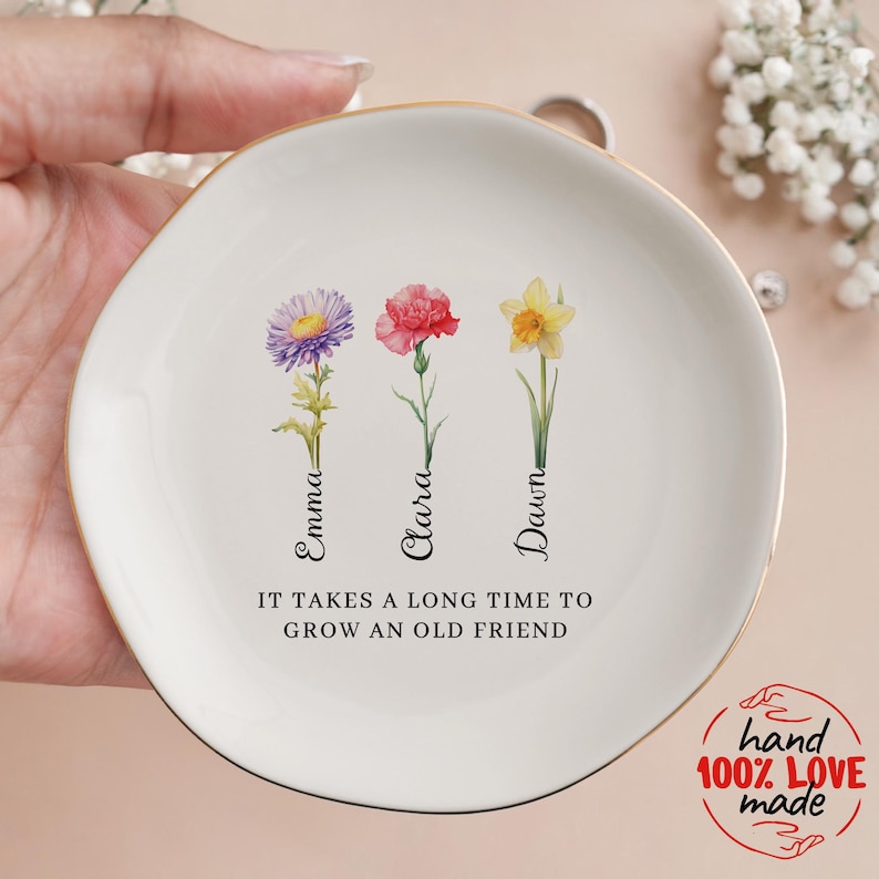 Pu&ograve; includere: Un piatto porta anelli in ceramica bianca con un bordo dorato. Presenta tre fiori ad acquerello con i nomi Emma, Clara e Dawn scritti sotto. Il piatto ha il testo "Ci vuole molto tempo per far crescere un vecchio amico".