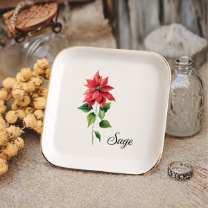 May include: A square, off-white ceramic tray with a gold rim. The tray features a red poinsettia flower with green leaves and the word "Sage" in script. A decorative ring and glass bottle are in the background.
