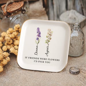 Pu&ograve; includere: Vassoio portaoggetti in ceramica bianca con bordo dorato, con un design floreale e il testo "Se gli amici fossero fiori, ti sceglierei". I fiori sono etichettati "Diana" e "Agnes".