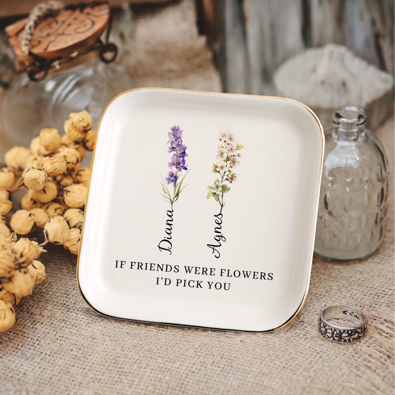 Pu&ograve; includere: Vassoio portaoggetti in ceramica bianca con bordo dorato, con un design floreale e il testo "Se gli amici fossero fiori, ti sceglierei". I fiori sono etichettati "Diana" e "Agnes".