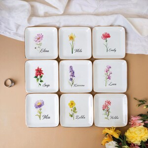 May include: Nine square white ceramic dishes with gold rims, each featuring a different watercolor floral design and a name. Flowers include pink, yellow, red, and purple blooms. Names include Elena, Mila, Emily, Sage, Katerina, Luca, Mia, Zelda, and Hilda.