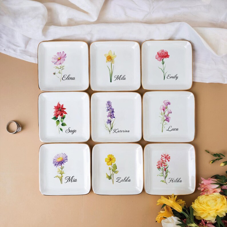 May include: Nine square white ceramic dishes with gold rims, each featuring a different watercolor floral design and a name. Flowers include pink, yellow, red, and purple blooms. Names include Elena, Mila, Emily, Sage, Katerina, Luca, Mia, Zelda, and Hilda.
