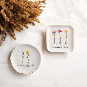 Pu&ograve; includere: Due piattini portaoggetti in ceramica bianca con bordi dorati. Il piattino pi&ugrave; piccolo ha un disegno di fiori di lavanda e gialli con il testo "Se gli amici fossero fiori, ti sceglierei". Il piattino pi&ugrave; grande ha tre fiori con il testo "Emma, Clara, Dawn" e "Ci vuole molto tempo per far crescere un vecchio amico."