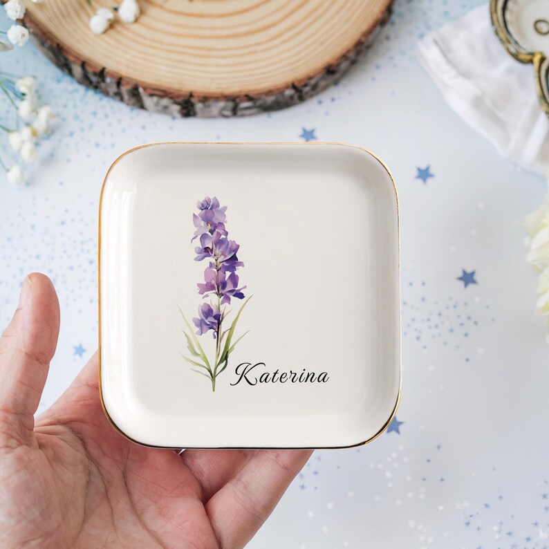 May include: A square, off-white ceramic trinket dish with a gold rim. It features a watercolor illustration of a purple flower and the name "Katerina" in black script. The dish is held in a hand.