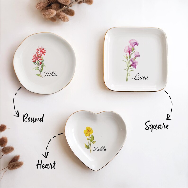 May include: Three white ceramic dishes with gold rims, each featuring a floral design and a name. The shapes include a round plate with red flowers and the name "Hilda", a square plate with purple flowers and the name "Luca", and a heart-shaped dish with a yellow flower and the name "Zelda".