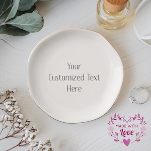 May include: White ceramic ring dish with a gold rim. The dish has the text "Your Customized Text Here" on it. The dish is surrounded by white flowers and a small bottle of perfume. The image also includes a pink heart with the text "Made with Love" in a floral wreath.