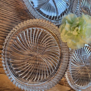 May include: Close-up view of four clear glass decorative plates. Each plate features a swirling, radial pattern radiating from the center. The plates are layered, creating a visually appealing display. A light green flower is positioned between the plates, adding a touch of color to the image. The plates appear to be made of high-quality, thick glass, with intricate detailing in the design.  The overall aesthetic is elegant and sophisticated, suggesting a use for formal settings or as decorative pieces.