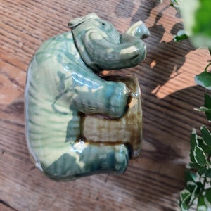 May include: A green ceramic elephant figurine with a textured surface. The elephant is sitting on a brown base.