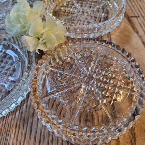 May include: Close-up view of several clear glass coasters. Each coaster features a diamond-patterned design and a slightly raised edge. The coasters are arranged on a wooden surface, partially overlapping.  The glass is transparent and reflects light, showcasing the intricate detailing of the cut glass.  These vintage-style coasters would be ideal for protecting furniture from drink spills.