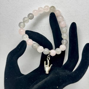 Può includere: Un braccialetto di perline grigie e rosa con un ciondolo d'argento a forma di mano che fa il segno 'rock on'.