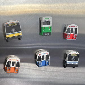 May include: A collection of colorful Boston transit train and bus magnets. The magnets feature various color schemes, including yellow, green, red, orange, blue, and purple. Each magnet has the word "BOSTON" on it.