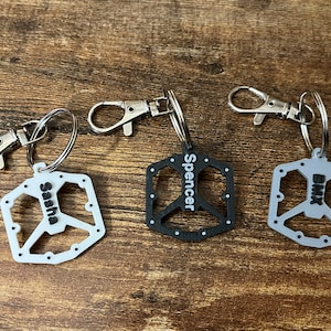 Puede incluir: Tres llaveros personalizados con un dise&ntilde;o de pedal de bicicleta. Uno es blanco con "Sasha" impreso, otro es negro con "Spencer", y el tercero es gris con "BMX". Cada llavero tiene un cierre plateado.