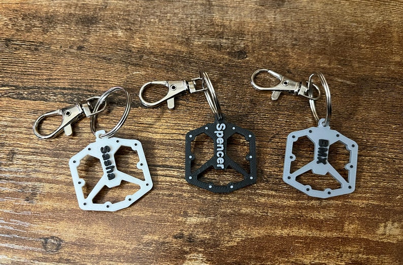 Puede incluir: Tres llaveros personalizados con un dise&ntilde;o de pedal de bicicleta. Uno es blanco con "Sasha" impreso, otro es negro con "Spencer", y el tercero es gris con "BMX". Cada llavero tiene un cierre plateado.
