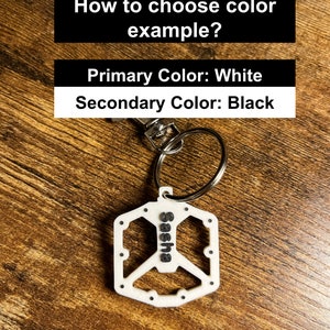 Puede incluir: Un llavero blanco con el nombre "Sasha" en negro y un dise&ntilde;o de pedal de bicicleta. El llavero est&aacute; sujeto a un llavero plateado. El texto superior dice "&iquest;C&oacute;mo elegir el ejemplo de color?" y "Color primario: Blanco, Color secundario: Negro."