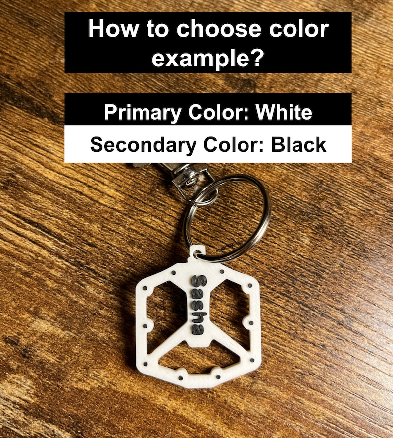 Puede incluir: Un llavero blanco con el nombre "Sasha" en negro y un dise&ntilde;o de pedal de bicicleta. El llavero est&aacute; sujeto a un llavero plateado. El texto superior dice "&iquest;C&oacute;mo elegir el ejemplo de color?" y "Color primario: Blanco, Color secundario: Negro."