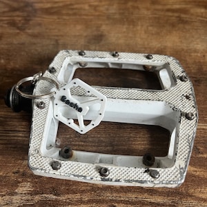 Puede incluir: Un pedal de bicicleta blanco con una superficie texturizada y pernos met&aacute;licos. Una peque&ntilde;a etiqueta blanca con el nombre "Sasha" est&aacute; sujeta al pedal con un llavero. El pedal est&aacute; sobre un fondo de madera.