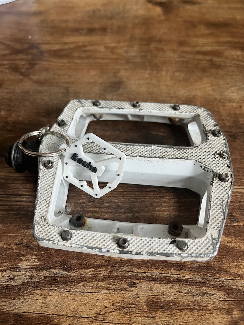 Puede incluir: Un pedal de bicicleta blanco con una superficie texturizada y pernos met&aacute;licos. Una peque&ntilde;a etiqueta blanca con el nombre "Sasha" est&aacute; sujeta al pedal con un llavero. El pedal est&aacute; sobre un fondo de madera.