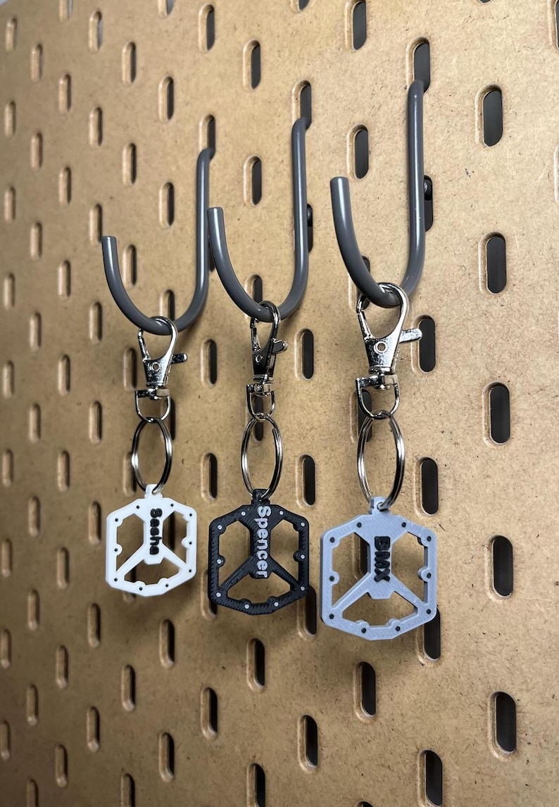 Puede incluir: Tres llaveros con dise&ntilde;o de pedal de bicicleta, cada uno con un color y nombre diferente. Los llaveros son blancos, negros y grises, y est&aacute;n colgados de un panel perforado. Los nombres "Sasha", "Spencer" y "BMX" est&aacute;n impresos en los llaveros.