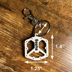 Puede incluir: Llavero blanco con el s&iacute;mbolo de la paz y el nombre "Sasha" en letras negras. El llavero mide 3,5 cm de largo, 3,2 cm de ancho y el anillo tiene un di&aacute;metro de 2,5 cm. Tiene un cierre y un anillo plateados.