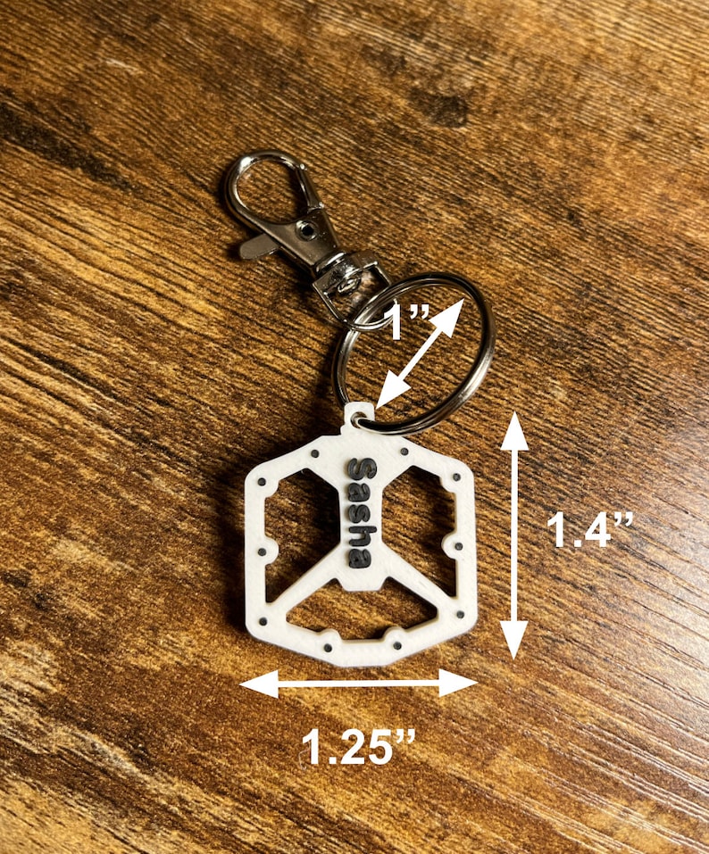 Puede incluir: Llavero blanco con el s&iacute;mbolo de la paz y el nombre "Sasha" en letras negras. El llavero mide 3,5 cm de largo, 3,2 cm de ancho y el anillo tiene un di&aacute;metro de 2,5 cm. Tiene un cierre y un anillo plateados.