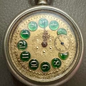 Può includere: Un orologio da tasca in argento con quadrante dorato e numeri in vetro verde. L'orologio presenta un design floreale decorativo e un piccolo quadrante secondario per i secondi.