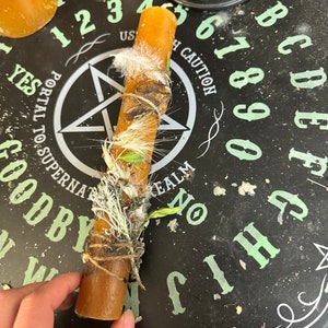 Könnte beinhalten: Eine braune Kerze mit getrockneten Kräutern und einer weißen Feder, die um sie herumgewickelt ist, auf einem schwarzen Ouija-Brett mit grüner Beschriftung. Das Brett enthält die Zahlen 1-9, 0 und die Buchstaben A-Z sowie die Wörter "Ja", "Nein", "Auf Wiedersehen", "Goodby", "Reich", "Zu Superna", "Portal zu Sonnen" und "Vorsicht".