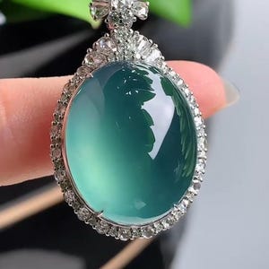 May include: A jade pendant necklace with a large, oval-shaped green jade stone surrounded by a halo of small diamonds. The pendant is attached to a black cord.