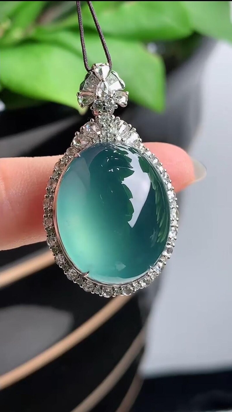May include: A jade pendant necklace with a large, oval-shaped green jade stone surrounded by a halo of small diamonds. The pendant is attached to a black cord.