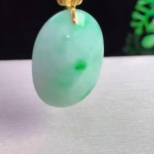 May include: Oval-shaped jade pendant with a light green hue and darker green spots. The pendant is suspended from a black cord with a gold-colored clasp. The jade has a smooth, polished surface.