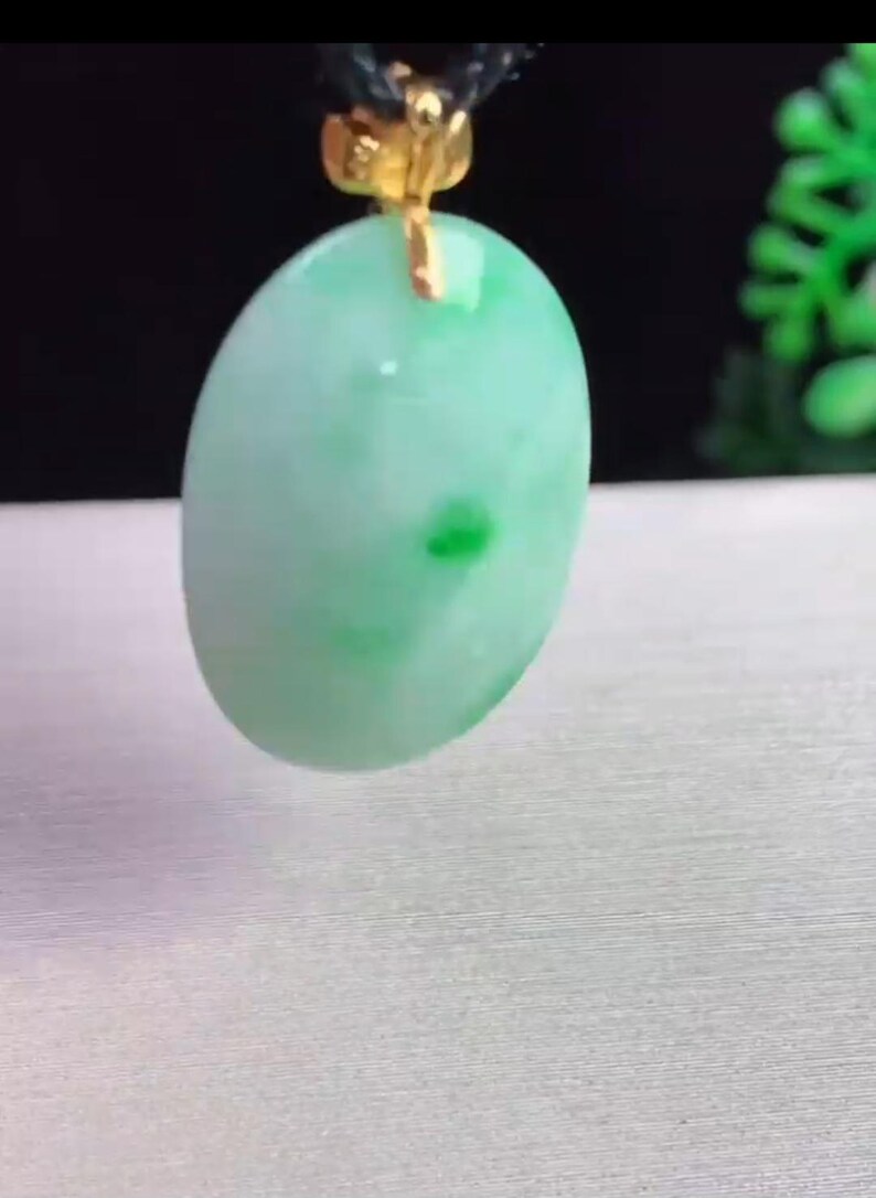 May include: Oval-shaped jade pendant with a light green hue and darker green spots. The pendant is suspended from a black cord with a gold-colored clasp. The jade has a smooth, polished surface.