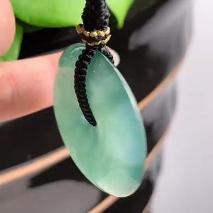 Pu&ograve; includere: Un ciondolo di giada verde con una forma ovale liscia appeso a un cordoncino nero. Il ciondolo &egrave; attaccato al cordoncino con un anello dorato.