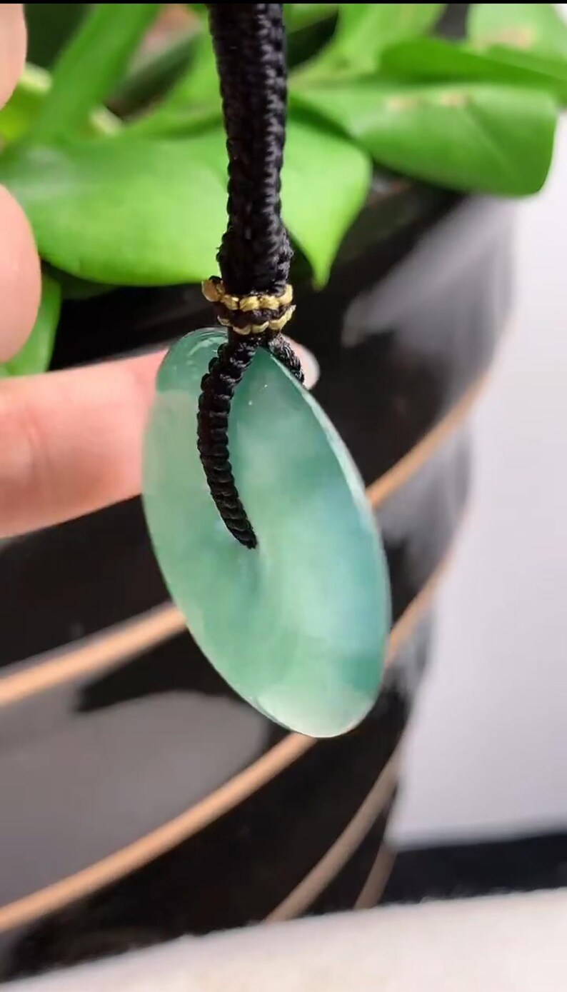 Pu&ograve; includere: Un ciondolo di giada verde con una forma ovale liscia appeso a un cordoncino nero. Il ciondolo &egrave; attaccato al cordoncino con un anello dorato.