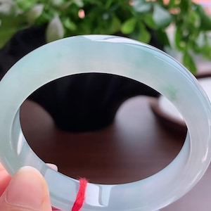 Può includere: Un bracciale rigido in giada verde pallido, liscio e traslucido, con sottili inclusioni bianche e verde scuro. La fascia circolare presenta una superficie lucida e arrotondata, accentuata da un sottile filo rosso legato su un lato.