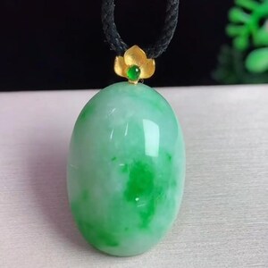 May include: A jade pendant with a green oval stone. The stone has lighter green patches and is attached to a black cord. A gold-colored flower-shaped bail with a small green stone accents the pendant.
