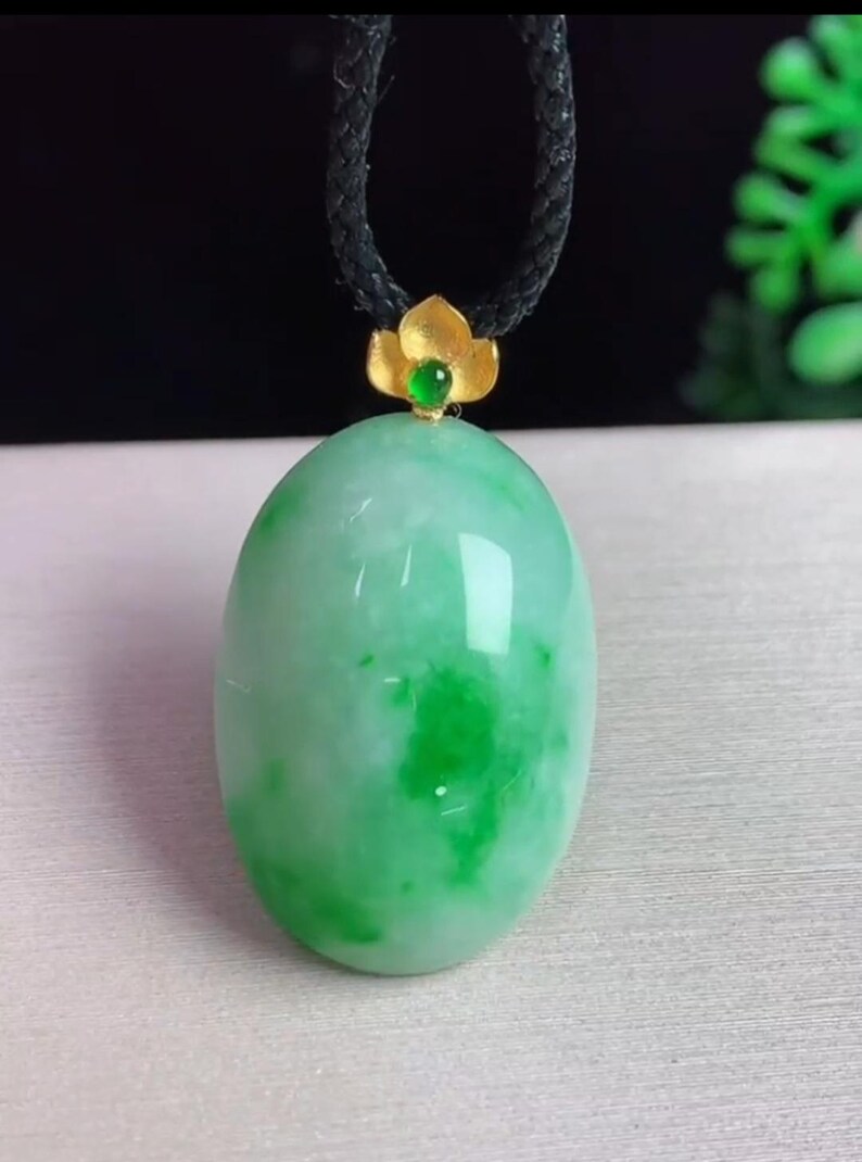 May include: A jade pendant with a green oval stone. The stone has lighter green patches and is attached to a black cord. A gold-colored flower-shaped bail with a small green stone accents the pendant.