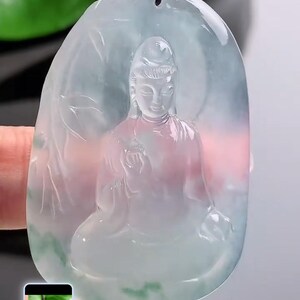 May include: A translucent jade pendant carved with a seated Buddha figure. The pendant is light green with pink and white hues, suspended from a black cord. The Buddha is depicted in a meditative pose.