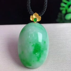 May include: A green jade pendant with a gold flower accent and a black cord. The oval-shaped jade has a marbled appearance with darker green patches. The gold accent holds a small green gem.