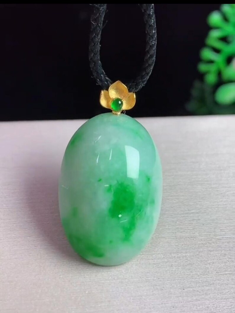 May include: A green jade pendant with a gold flower accent and a black cord. The oval-shaped jade has a marbled appearance with darker green patches. The gold accent holds a small green gem.