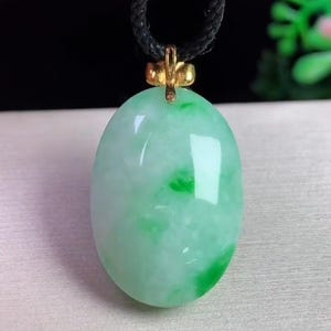 May include: Oval jade pendant with a gold-colored bail, suspended from a black cord. The jade is a light green color with darker green veining. The pendant is smooth and polished, reflecting light.