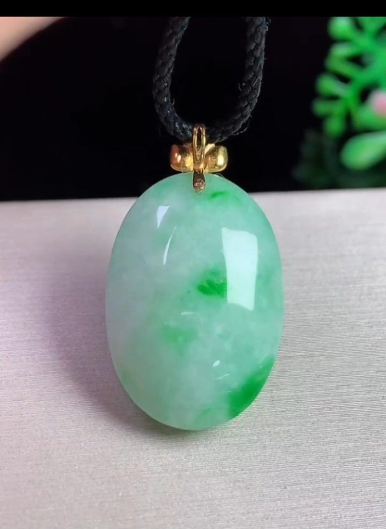 May include: Oval jade pendant with a gold-colored bail, suspended from a black cord. The jade is a light green color with darker green veining. The pendant is smooth and polished, reflecting light.