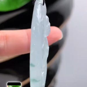 May include: A pale green jade pendant with a carved figure, hanging from a black cord. The jade displays subtle colour variations and is set against a blurred backdrop of green leaves.