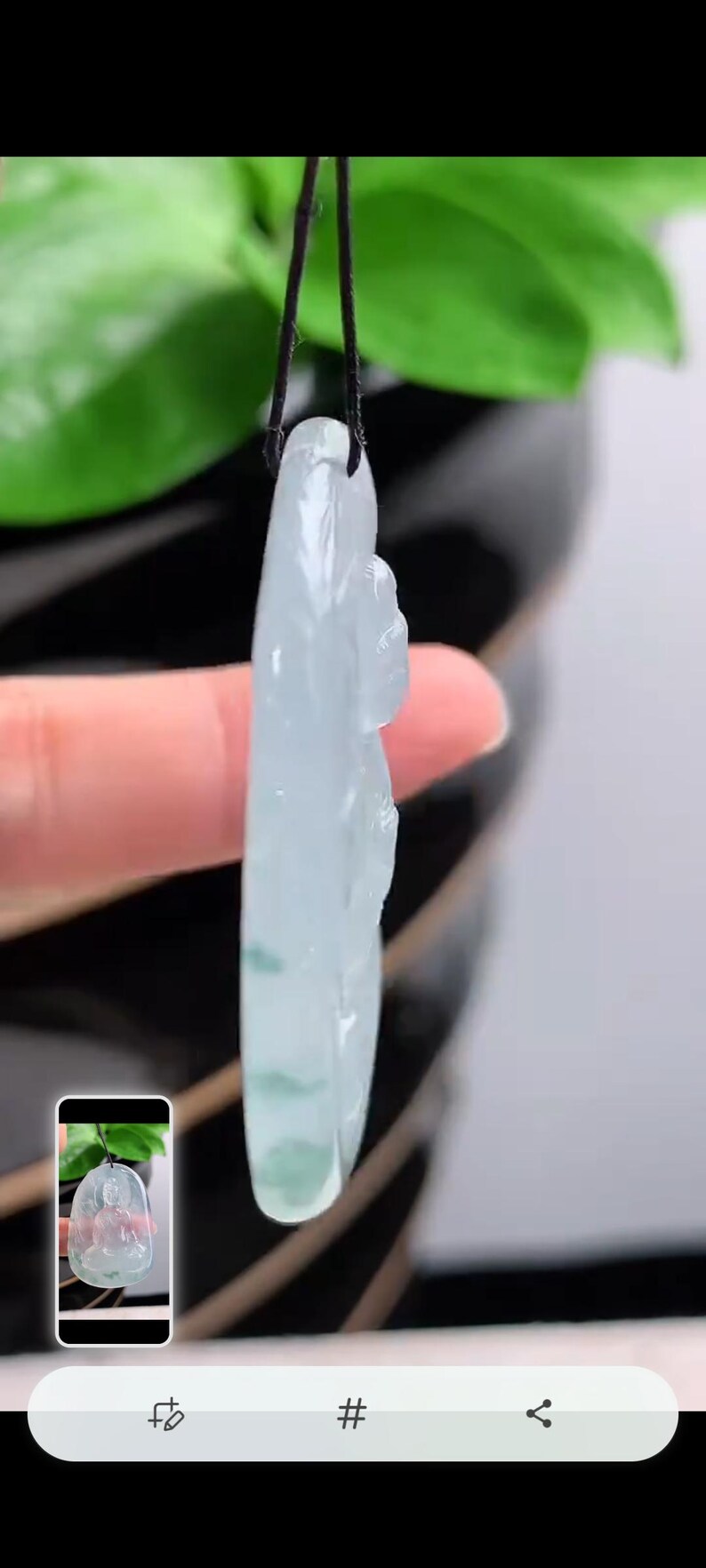 May include: A pale green jade pendant with a carved figure, hanging from a black cord. The jade displays subtle colour variations and is set against a blurred backdrop of green leaves.