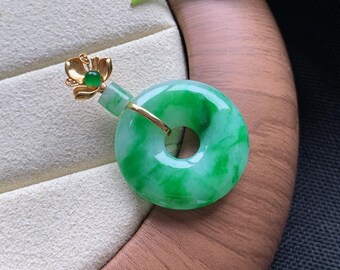May include: A circular green jade pendant featuring a gold-toned metal bail shaped like a flower with a small green stone at its center. The jade ring has a smooth, polished surface with natural variations in color, ranging from light to deep green.