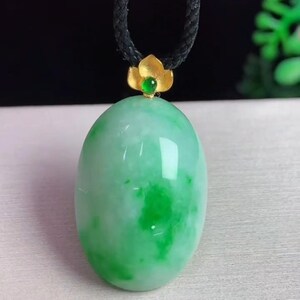 May include: A jade pendant necklace featuring a large, oval-shaped, light green jade stone. The jade has darker green marbling. The pendant is topped with a gold-colored flower and a small green gemstone, suspended from a black cord.