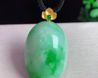 May include: A jade pendant necklace featuring a large, oval-shaped, light green jade stone. The jade has darker green marbling. The pendant is topped with a gold-colored flower and a small green gemstone, suspended from a black cord.